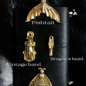 May include: A collection of gold-colored metal decorative objects, including a fishtail, a vintage hand, a dragon's head, and a gold moth. The objects are displayed on a black leather surface. Text labels identify each object.