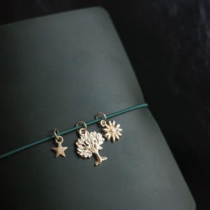 May include: A dark green journal with a gold star, tree, and sun charm bracelet. The charms are attached to a green elastic band. The journal has a smooth, matte finish and is set against a dark background.