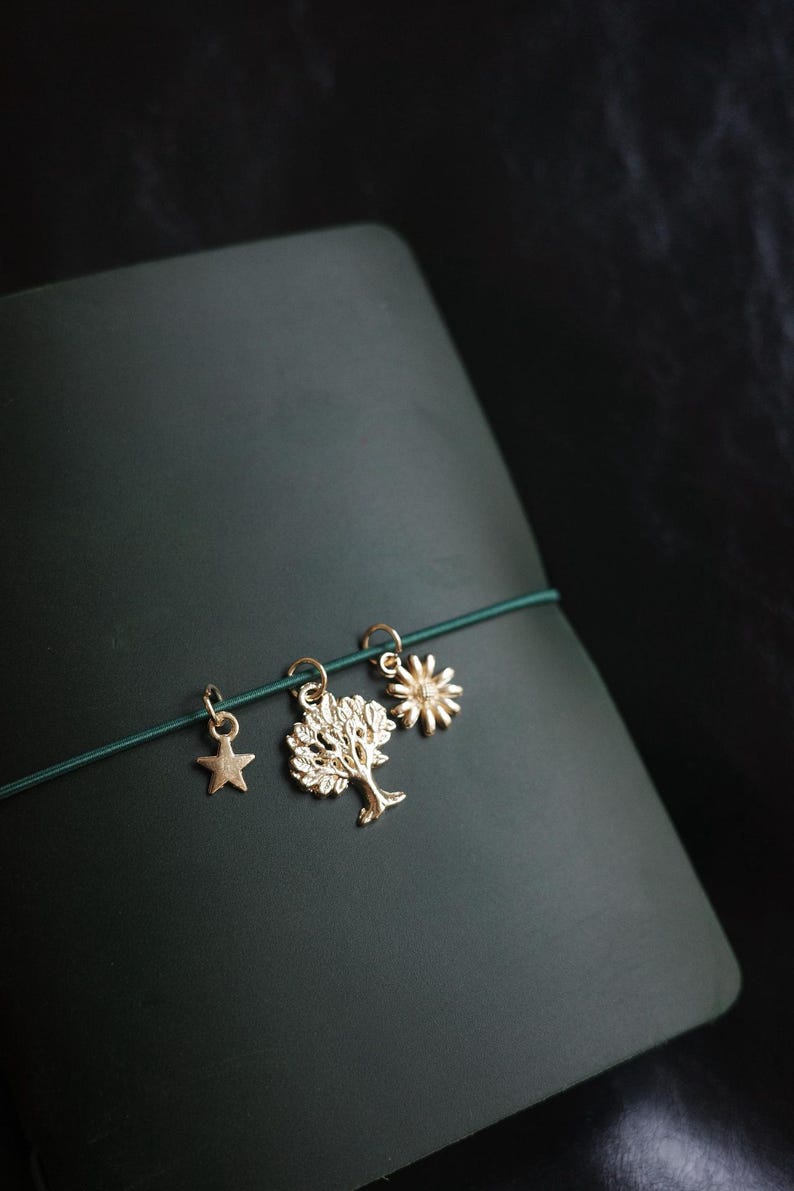 May include: A dark green journal with a gold star, tree, and sun charm bracelet. The charms are attached to a green elastic band. The journal has a smooth, matte finish and is set against a dark background.