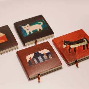 Puede incluir: Cuatro cuadernos cuadrados con portadas pintadas. Uno muestra un gato blanco sobre fondo oscuro, otro un burro sobre fondo rojo, otro un paisaje de montaña sobre fondo marrón y otro una forma naranja parcial sobre fondo marrón oscuro.