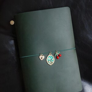 May include: Dark green leather journal with a decorative elastic band. The band holds three charms: a heart-shaped lock, an oval pendant with a floral design, and a pair of red cherries.