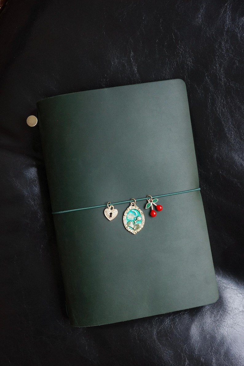 May include: Dark green leather journal with a decorative elastic band. The band holds three charms: a heart-shaped lock, an oval pendant with a floral design, and a pair of red cherries.