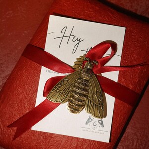 May include: A red gift box wrapped with a red ribbon, adorned with a gold-colored bee brooch and a white card that says "Hey". The bee brooch is detailed with intricate wing and body patterns. The gift box has a textured surface.