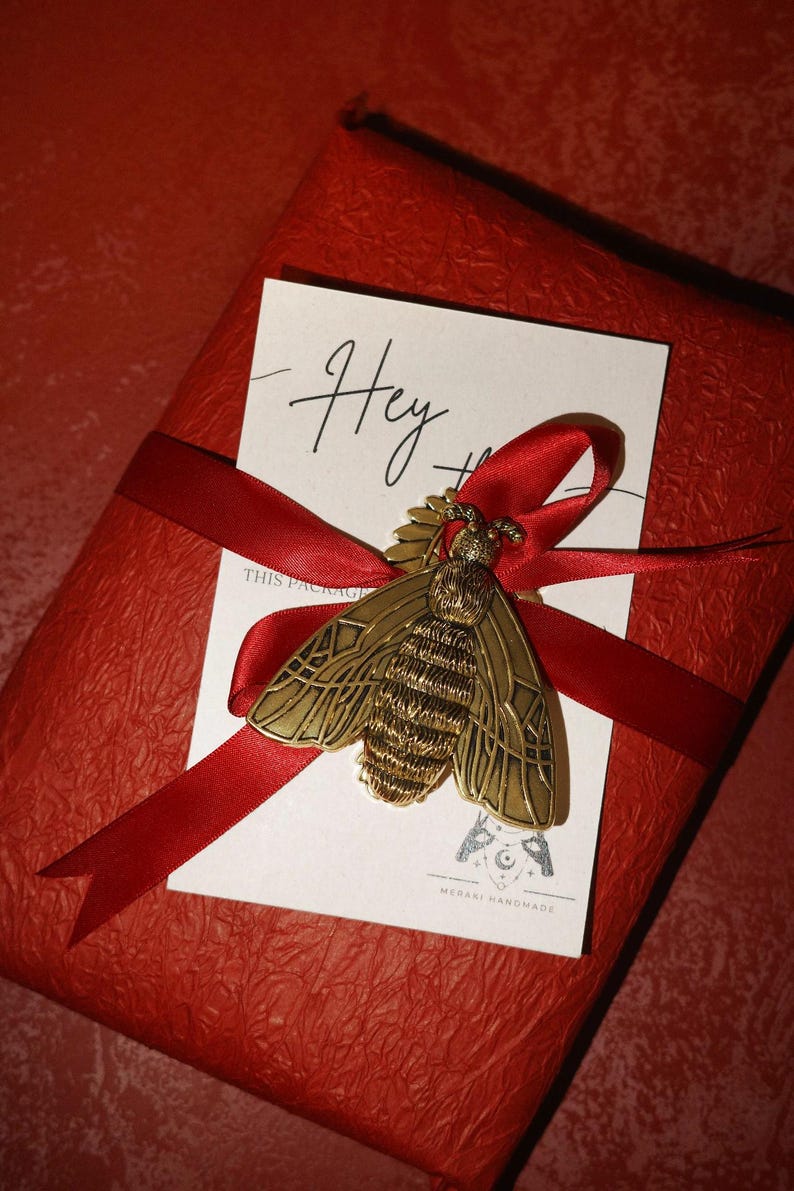 May include: A red gift box wrapped with a red ribbon, adorned with a gold-colored bee brooch and a white card that says "Hey". The bee brooch is detailed with intricate wing and body patterns. The gift box has a textured surface.