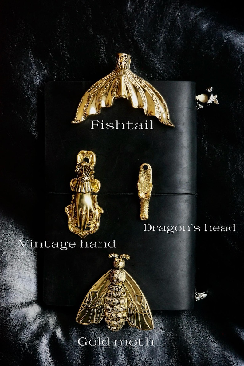 May include: A collection of gold-colored metal decorative objects, including a fishtail, a vintage hand, a dragon's head, and a gold moth. The objects are displayed on a black leather surface. Text labels identify each object.