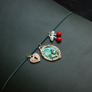May include: A collection of gold-toned charms on a green cord. The charms include a heart with a keyhole, a floral oval pendant, and a pair of red cherry charms. The background is a dark green surface.