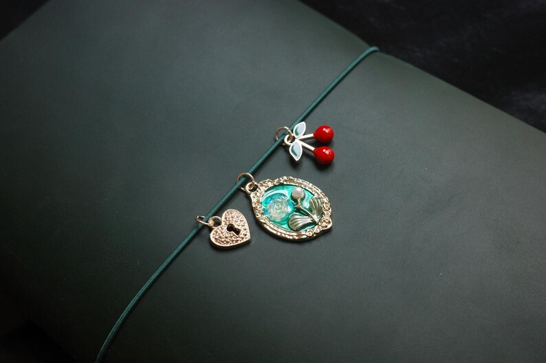 May include: A collection of gold-toned charms on a green cord. The charms include a heart with a keyhole, a floral oval pendant, and a pair of red cherry charms. The background is a dark green surface.