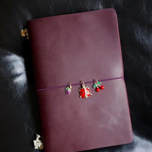 May include: A burgundy leather journal with a purple elastic closure. The journal is adorned with three decorative charms: a purple grape cluster, a red ladybug, and a pair of red cherries. The journal is on a black leather surface.