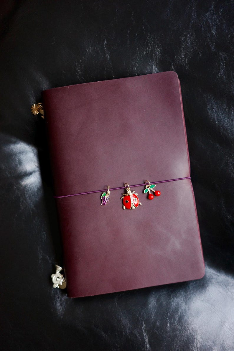 May include: A burgundy leather journal with a purple elastic closure. The journal is adorned with three decorative charms: a purple grape cluster, a red ladybug, and a pair of red cherries. The journal is on a black leather surface.