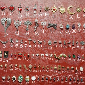 May include: A collection of small, decorative charms in various shapes and designs, including hearts, crosses, stars, and moons. The charms are arranged on a red surface and labeled with letters and numbers.