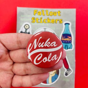 Op de afbeelding: Een rode Nuka Cola-knop met witte tekst wordt in een hand gehouden. De knop maakt deel uit van een set Fallout-stickers, met andere stickerontwerpen zichtbaar op de achtergrond. De achtergrond is rood.