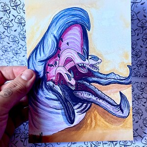 May include: A rectangular art print featuring a colorful illustration of a creature with a large blue head, a pink open mouth, and multiple smaller tentacle-like appendages with sharp teeth extending outward against a warm yellow background.