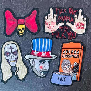 Op de afbeelding: Een verzameling stickers met diverse ontwerpen. Eén toont een rode strik met een schedel, een ander toont handen met aanstootgevende tekst. Andere stickers zijn een zombie, een man met een patriottische hoed en een ontbijtgranendoos met het woord "TINY".