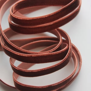 May include: A close-up of a spool of rust-colored velvet ribbon. The ribbon is soft and has a subtle sheen.
