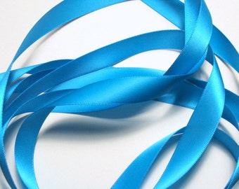 May include: Bright turquoise satin ribbon, arranged in looping curves against a white background. The ribbon has a smooth, glossy texture and a slight sheen, suggesting it is made of a high-quality material. The image is well-lit, highlighting the ribbon's color and texture.