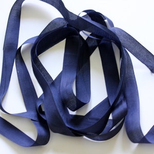 May include: Navy blue bias tape ribbon, arranged in a loose, looping pattern against a white background. The ribbon appears to be made of a woven fabric, with a subtle sheen. Ideal for sewing and crafting projects.