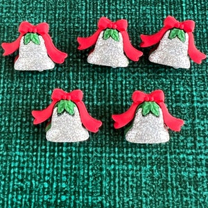 May include: Five Christmas bell ornaments. Each bell is silver with red bows and ribbons. Green holly leaves are at the top of each bell. The ornaments are on a textured green background.