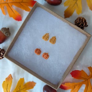 May include: A pair of small, orange pumpkin-shaped stud earrings and a pair of golden-yellow leaf-shaped stud earrings. The four earrings are arranged in a square formation inside a square cardboard gift box lined with white tissue paper.