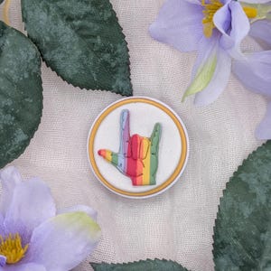 May include: A round, white and gold-edged charm featuring a rainbow-colored hand gesture, with the fingers forming the sign for "I love you". The charm is surrounded by green leaves and purple flowers.