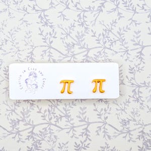 May include: Gold-colored pi symbol earrings on a white card with the text "Crafted in Clay and Chaos". The earrings are displayed on a floral patterned background. The earrings are a fun accessory for math enthusiasts.