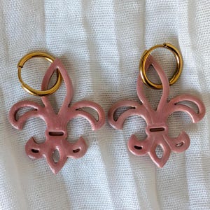 May include: A pair of pink fleur-de-lis earrings with gold-colored hoops. The earrings are a light pink color and have a glossy finish. The earrings are displayed on a white, textured surface.