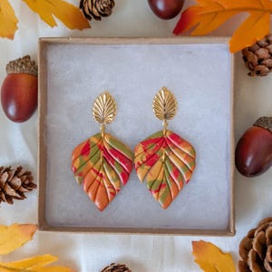 May include: A pair of dangle earrings featuring gold-tone metal leaf-shaped studs attached to larger, textured leaf-shaped pendants. The pendants display a marbled pattern of autumn colors including orange, red, yellow, and green tones.