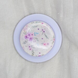May include: A circular button featuring a white background with a delicate watercolor pattern of purple and pink flowers, green leaves, and thin stems. The button is set within a smooth, light lavender circular frame with a raised, rounded edge.