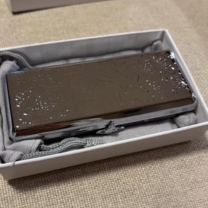 May include: A rectangular, metallic brown case with a decorative pattern, resting inside a white box with a gray fabric lining. The case has a silver clasp and is presented on a neutral-toned surface.