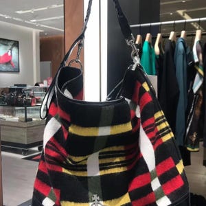 May include: A black, red, yellow, and white plaid shoulder bag with a black leather base and strap. The bag features a silver emblem and a braided handle. The bag is hanging in a retail store.