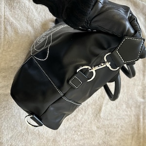 May include: Black handbag with white stitching and a floral design. The bag features a detachable shoulder strap with silver-tone hardware. The handles are black with white stitching. The bag is held by a gloved hand.
