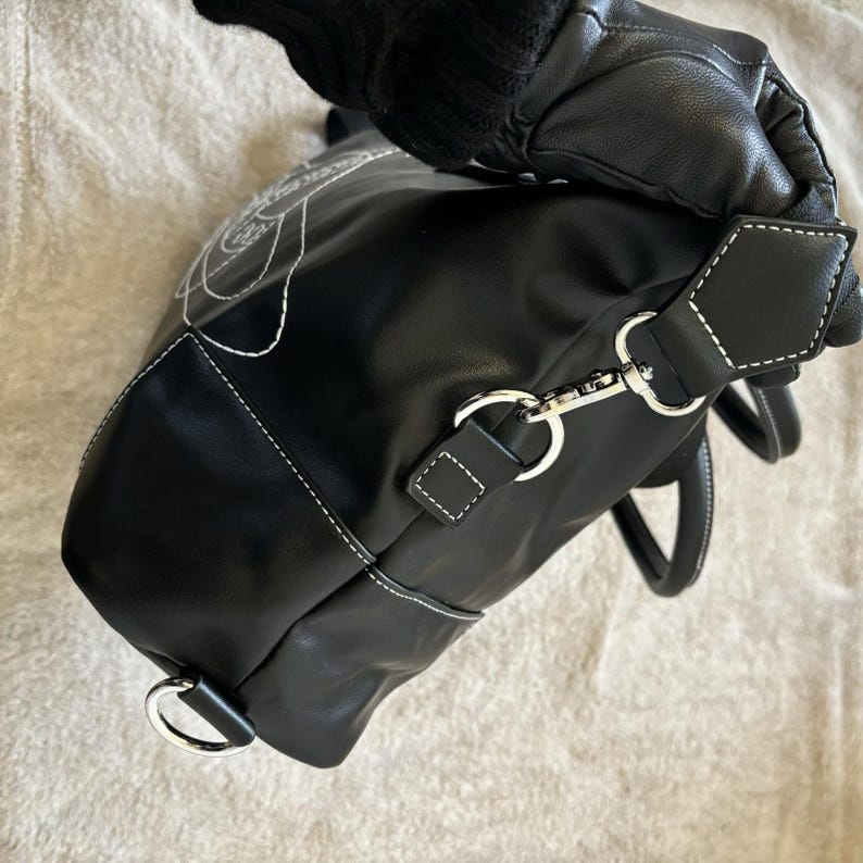 May include: Black handbag with white stitching and a floral design. The bag features a detachable shoulder strap with silver-tone hardware. The handles are black with white stitching. The bag is held by a gloved hand.