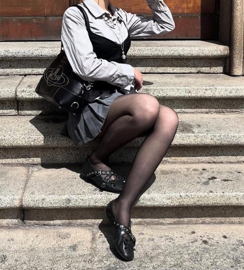 May include: A black leather shoulder bag with a white logo, a gray pleated skirt, and black shoes with silver accents. The outfit is completed with a white collared shirt and black tights. The person is sitting on stone steps.