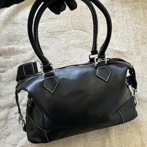 May include: Black leather handbag with white stitching details. Features two curved top handles and a detachable shoulder strap. Silver-tone hardware accents the bag's design. The bag has a zippered closure and a structured shape.