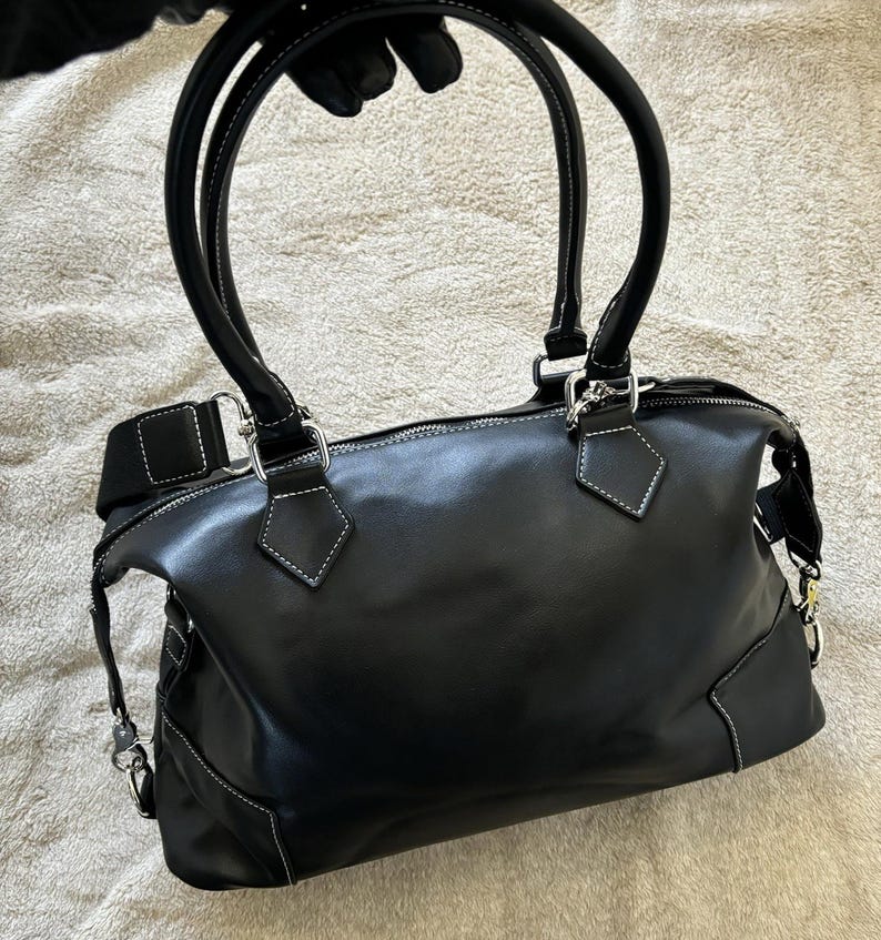 May include: Black leather handbag with white stitching details. Features two curved top handles and a detachable shoulder strap. Silver-tone hardware accents the bag's design. The bag has a zippered closure and a structured shape.