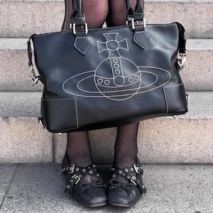 May include: Black leather handbag with white stitching and a planet design. The bag has two handles and a shoulder strap. The image also shows black shoes with buckles and bows.