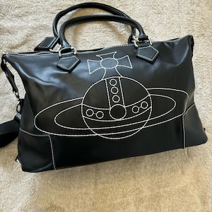 May include: Black tote bag with white stitching and a large, stylized planet design. The bag has two top handles, a shoulder strap, and a zipper closure. The bag is made of a smooth material.