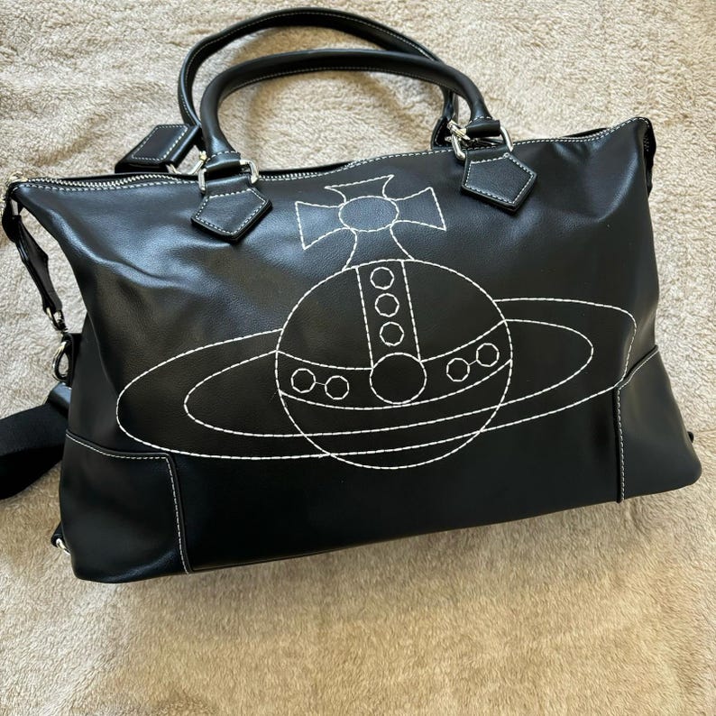 May include: Black tote bag with white stitching and a large, stylized planet design. The bag has two top handles, a shoulder strap, and a zipper closure. The bag is made of a smooth material.