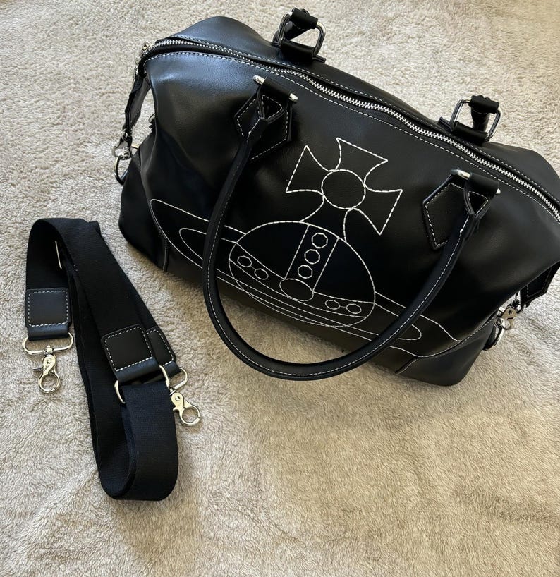 May include: Black handbag with white stitching and a white emblem. The bag has a zipper closure, two handles, and a detachable black shoulder strap with silver-tone hardware. The bag is made of a smooth material.