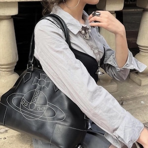 May include: Black leather handbag with white stitching featuring a planet design. The bag has a structured shape with top handles. The design is a stylized planet with a cross and circles. The bag is carried over the shoulder.