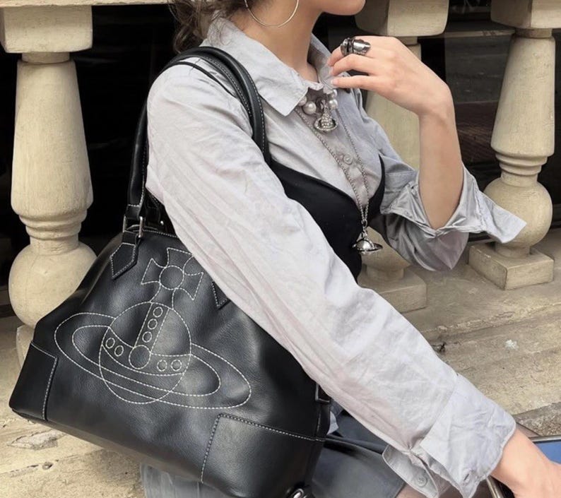 May include: Black leather handbag with white stitching featuring a planet design. The bag has a structured shape with top handles. The design is a stylized planet with a cross and circles. The bag is carried over the shoulder.