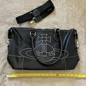 May include: Black handbag with white stitching and a white embroidered design. The bag has a black shoulder strap and silver-tone hardware. A measuring tape shows the bag is approximately 12 inches wide.