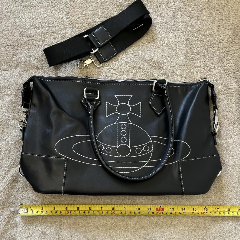 May include: Black handbag with white stitching and a white embroidered design. The bag has a black shoulder strap and silver-tone hardware. A measuring tape shows the bag is approximately 12 inches wide.