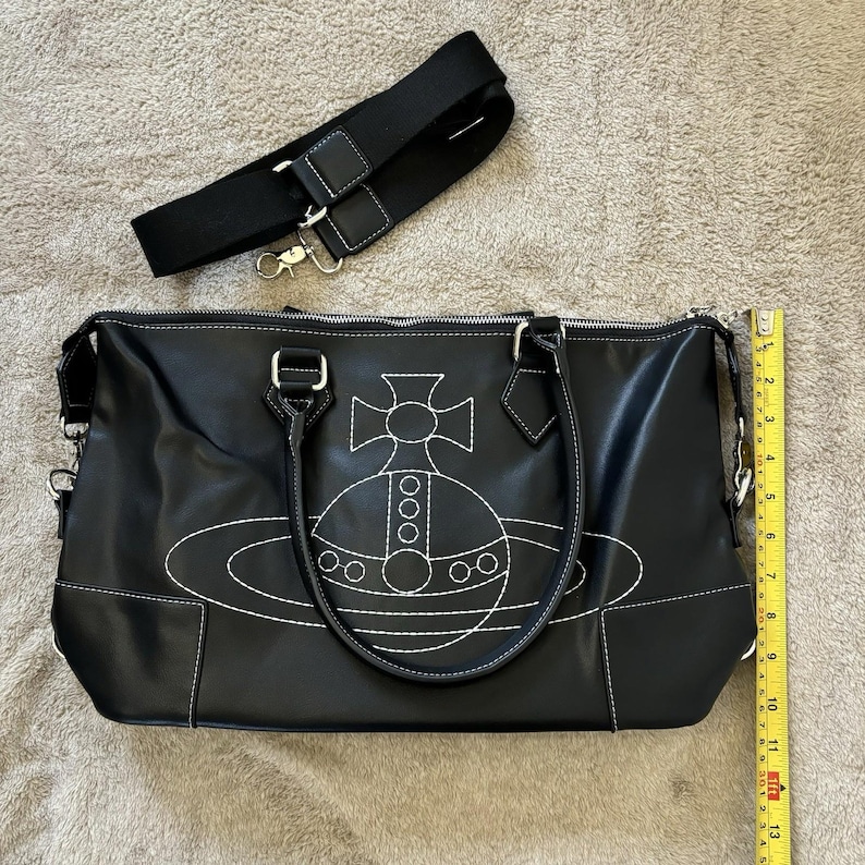 May include: Black handbag with white stitching and a white embroidered design. Includes a detachable black shoulder strap. The bag has two handles and a zipper closure. The bag is approximately 12 inches long.