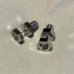 Puede incluir: Pendientes de tuerca plateados. Las cabezas hexagonales presentan un diseño gráfico negro. Los postes roscados y las cabezas hexagonales son visibles. Los pendientes se muestran sobre una tela gris claro.