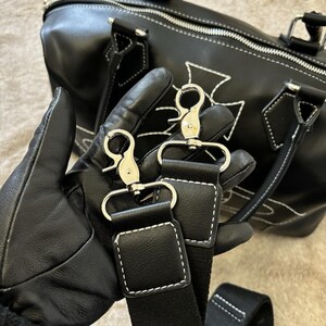 May include: Black leather duffel bag with white stitching and silver-tone hardware. The bag features a cross design and a detachable shoulder strap. The bag is held by a person wearing black gloves.