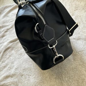 May include: Black duffel bag with white stitching and silver-tone hardware. The bag has a zipper closure and a detachable shoulder strap. The bag is made of a shiny material.