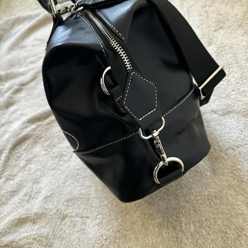 May include: Black duffel bag with white stitching and silver-tone hardware. The bag has a zipper closure and a detachable shoulder strap. The bag is made of a shiny material.