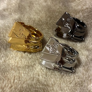 May include: Three rings in gold, silver, and gunmetal. Each ring features a layered design with a distinctive emblem. The rings are displayed on a textured, light-colored surface, showcasing their metallic shine.