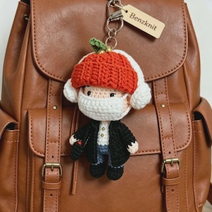 Può includere: Un portachiavi a forma di bambola all'uncinetto che raffigura un personaggio con un berretto arancione brillante, paraorecchie bianchi e una mascherina bianca. Il personaggio indossa una giacca nera sopra una camicia bianca e pantaloni blu. È attaccata una piccola etichetta in legno con la scritta Benzknit.
