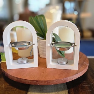 May include: Two white, arched candle holders with a silver dish and a small tea light candle. The holders sit on a round, wooden tray. Greenery and candles are in the background, creating a warm, inviting ambiance.